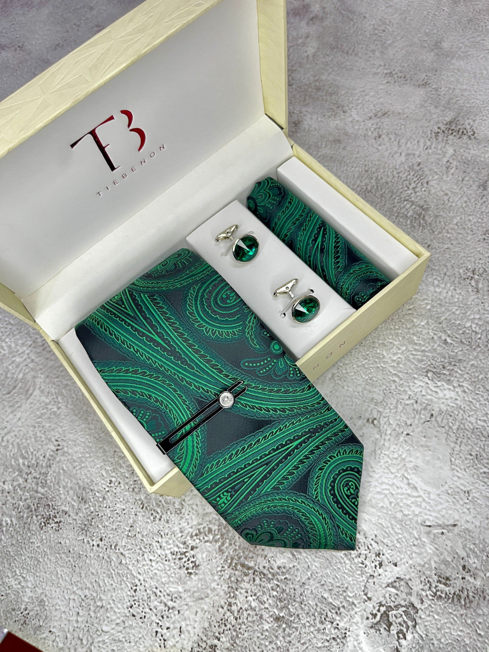 A set of two green paisley microsilk neckties with a patterned pocket square, displayed in an open white box with a gold interior, bearing the initial 'T' on the top of the box lid.