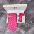 A hot pink paisley print silk tie with a brass tie pin, cufflinks, a lapel flower pin, and a matching pocket square, all presented in an open white box with a monogram 'TB' on the top and a pink rosebud on the side.