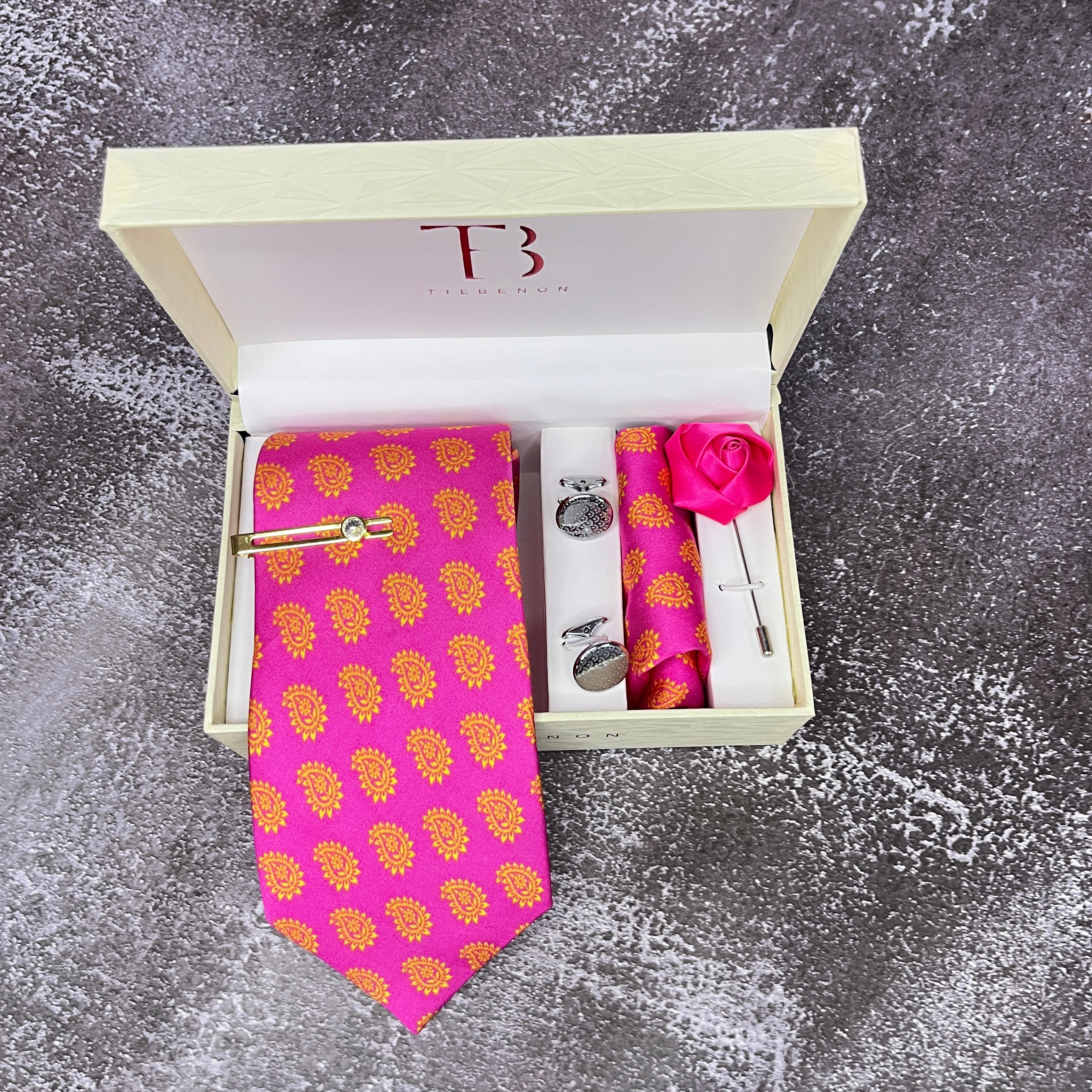 A hot pink paisley print silk tie with a brass tie pin, cufflinks, a lapel flower pin, and a matching pocket square, all presented in an open white box with a monogram 'TB' on the top and a pink rosebud on the side.