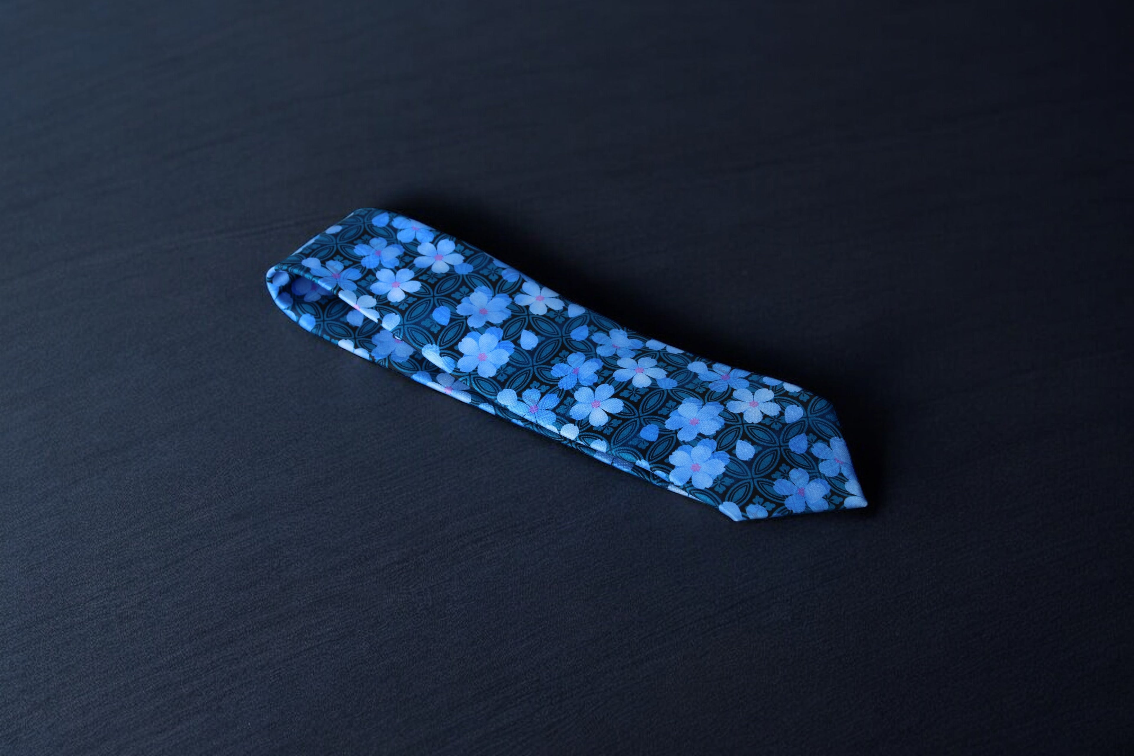 A blue necktie with a geometric pattern laid out flat on a dark background.