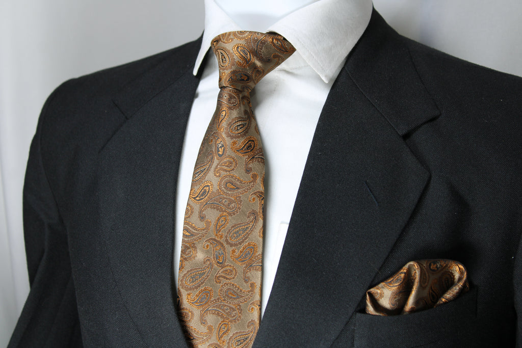 A light brown and gold paisley necktie paired with a matching pocket square displayed on a mannequin wearing a black suit and a white shirt.