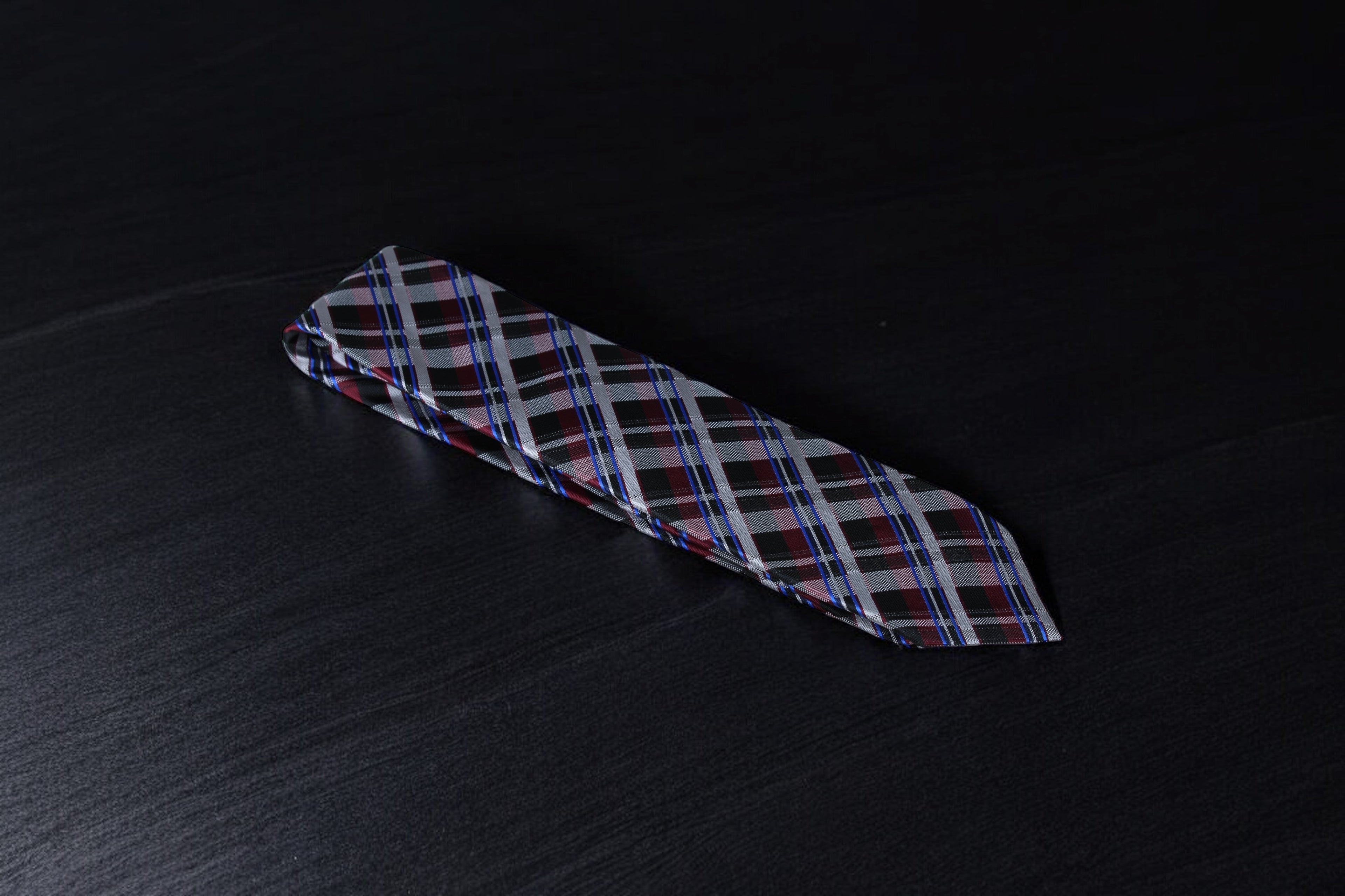 A checkered tie with maroon and black colors lying flat on a dark surface.