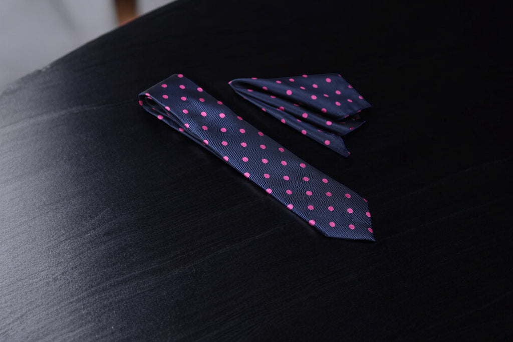 A navy and pink polka dotted tie and pocket square set laid out on a dark surface.