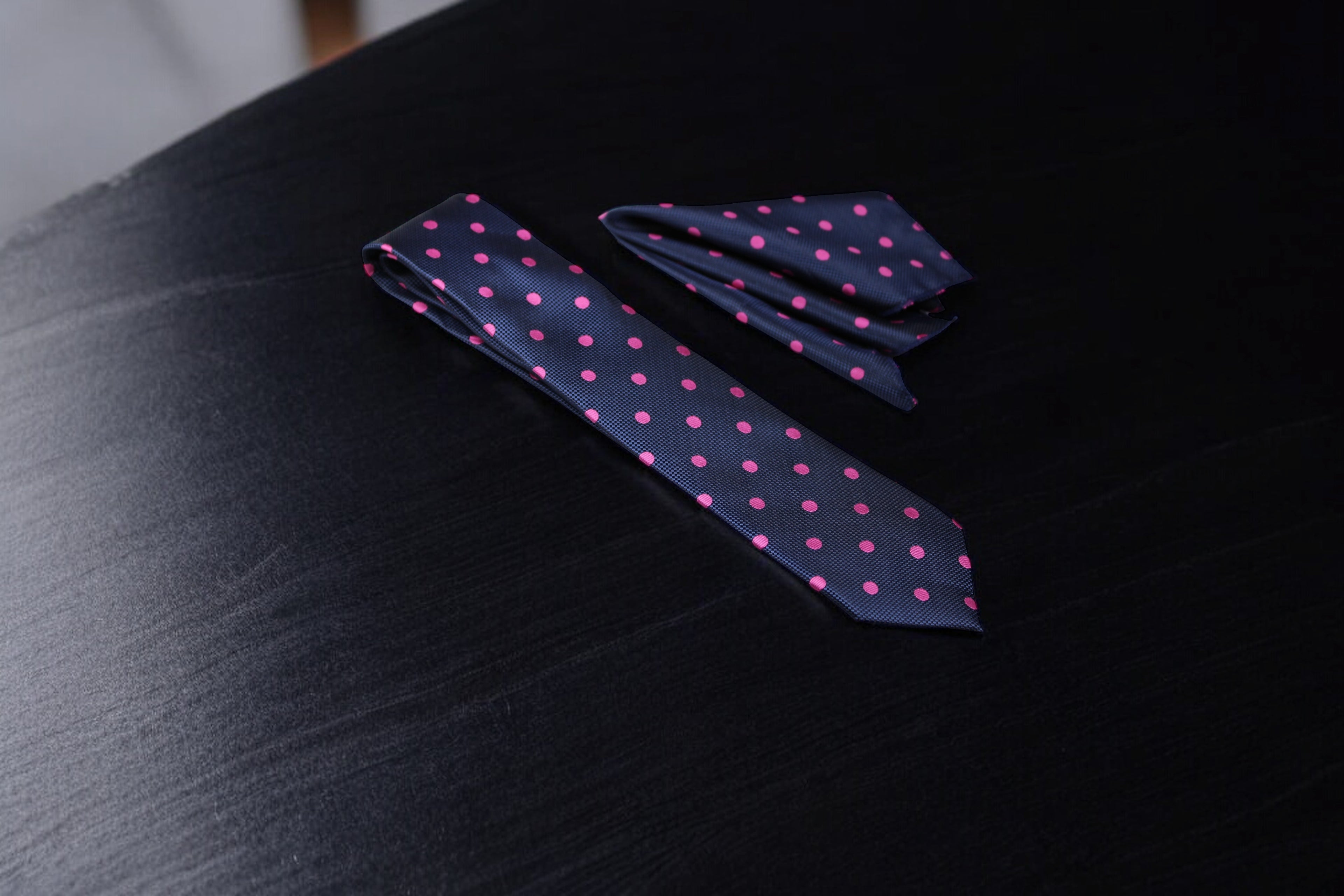 A navy and pink polka dotted tie and pocket square set laid out on a dark surface.