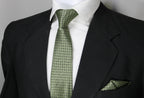 A set consisting of an olive green tie with a geometric pattern and a matching pocket square, displayed on a mannequin wearing a black suit and a white shirt.