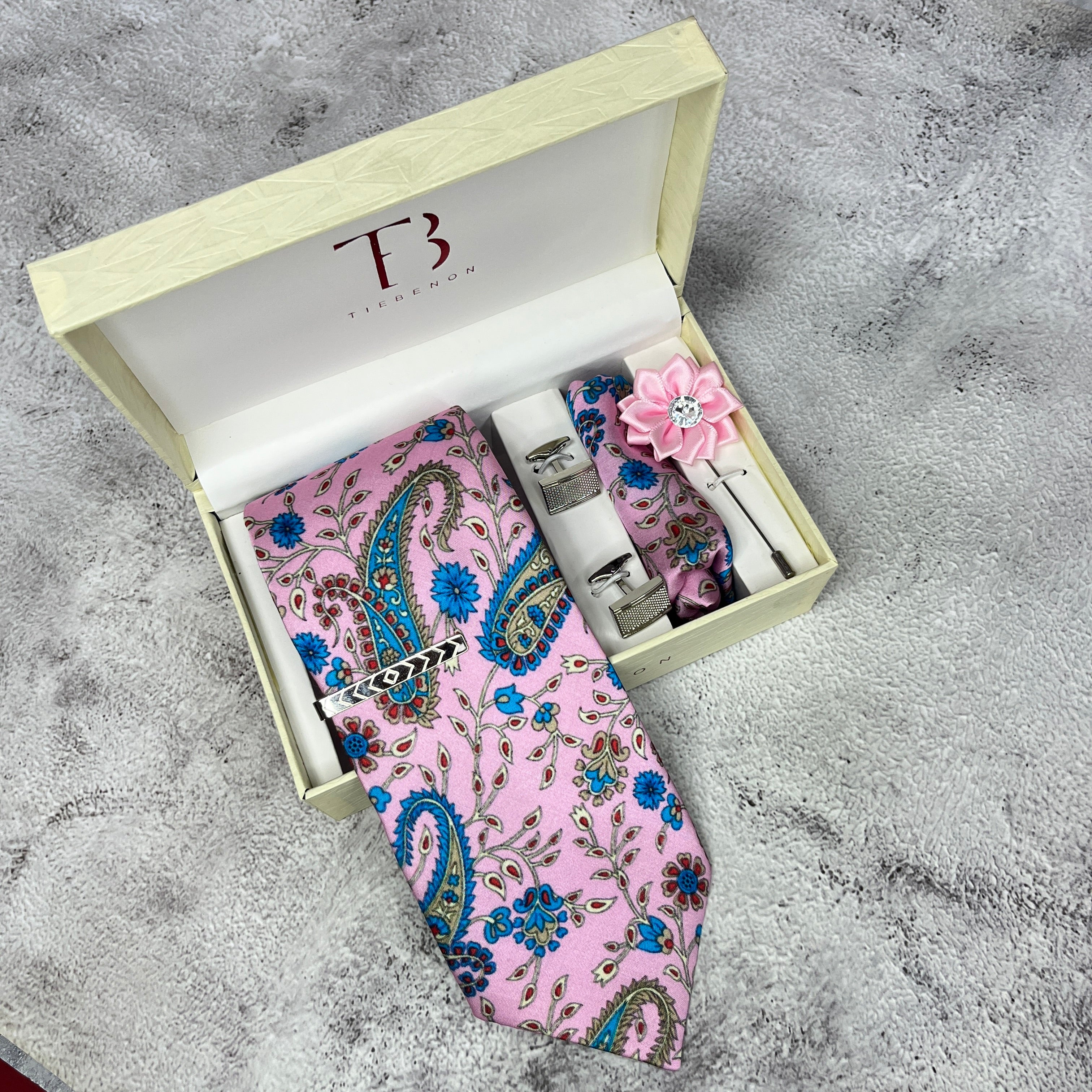 A pink paisley printed silk tie is displayed in an open white box with a branded 'T' on the top and a small floral decoration included.
