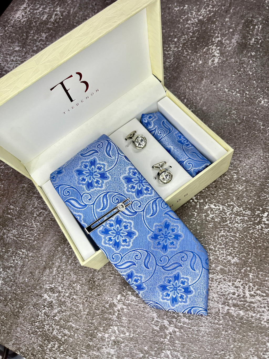 A set of men's formal wear including a blue microsilk tie with a floral pattern, a matching pocket square, and cufflinks, presented in an open white box with a TCHT logo on the top.