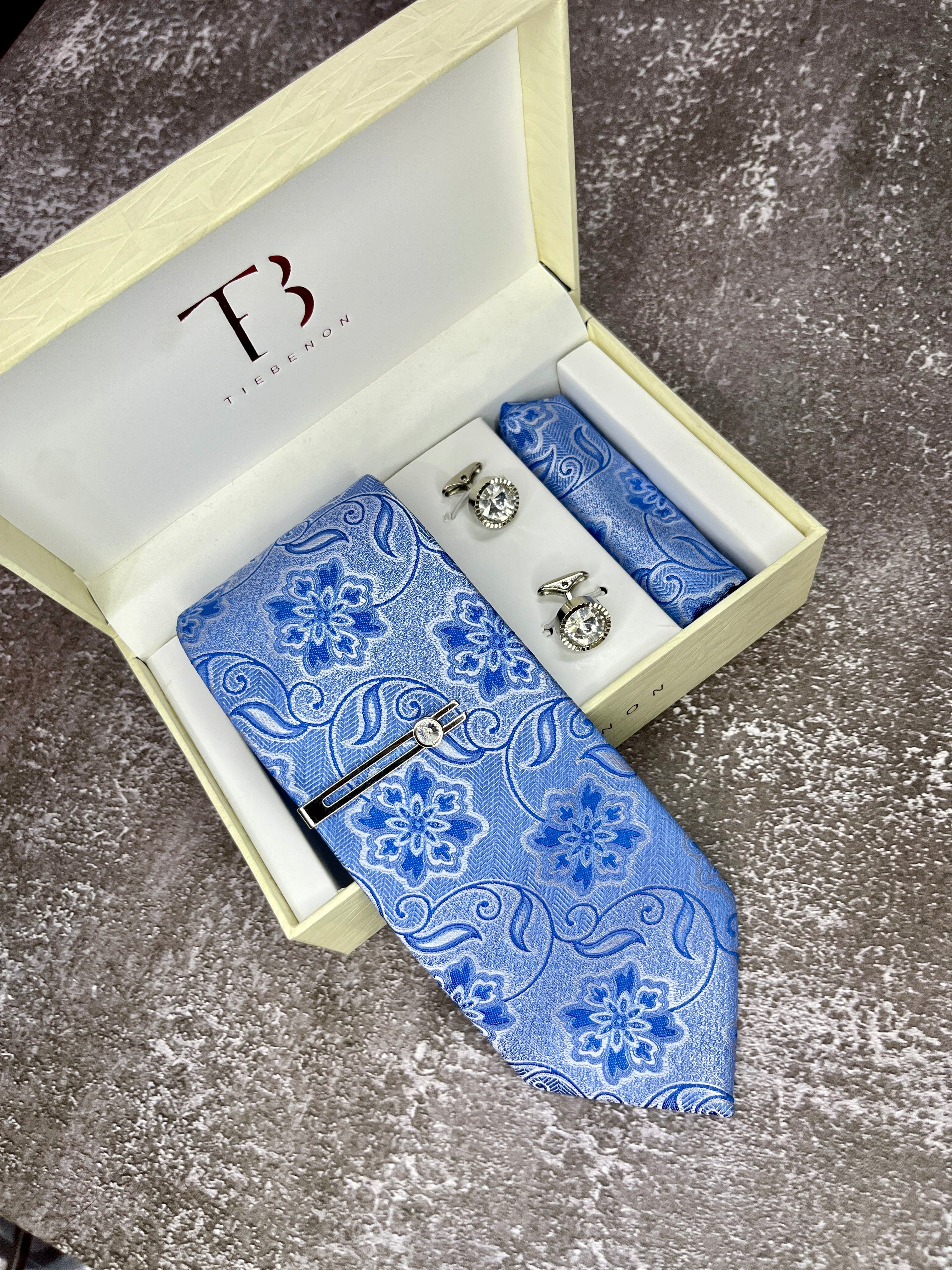 A set of men's formal wear including a blue microsilk tie with a floral pattern, a matching pocket square, and cufflinks, presented in an open white box with a TCHT logo on the top.