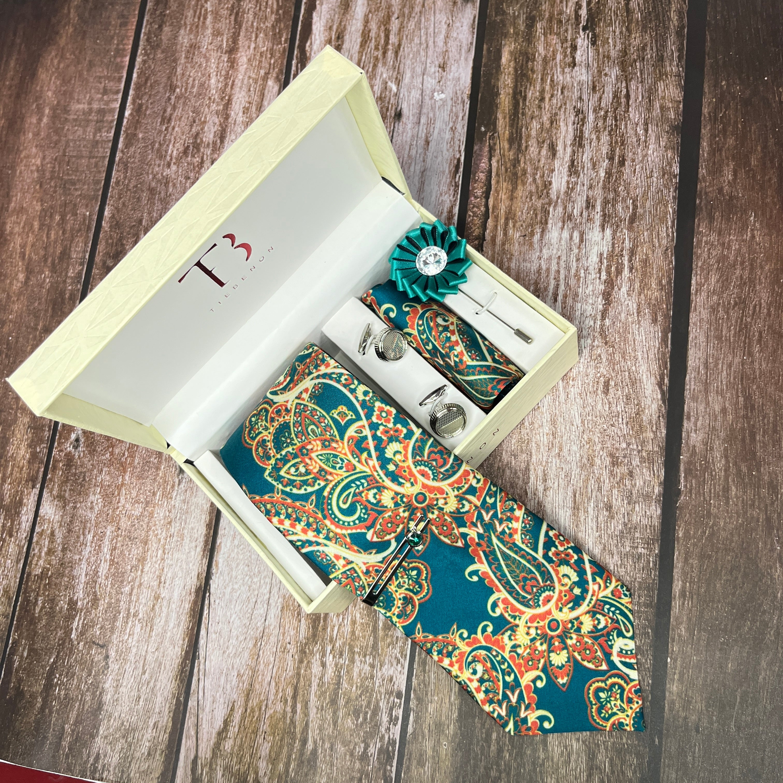 A teal paisley printed silk tie with a subtle paisley pattern, displayed in an open white box with a branded 'T' on the top and a green decorative flower pin.