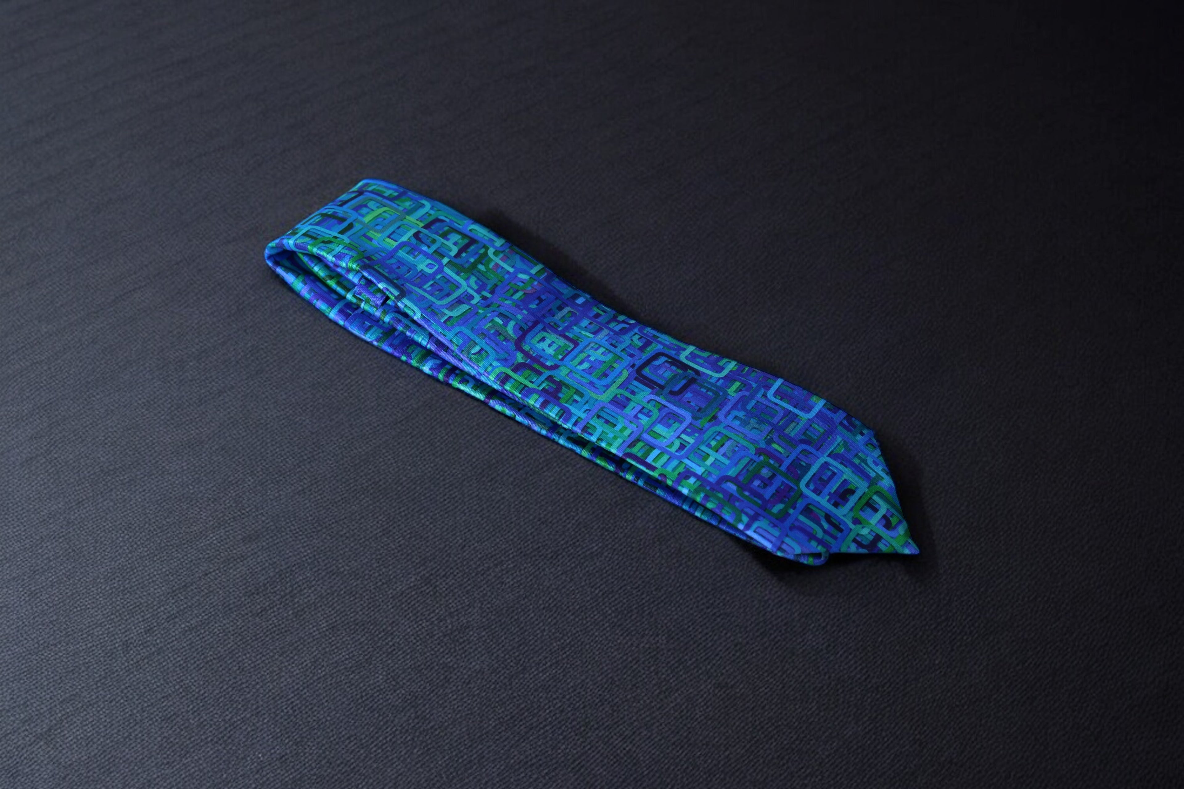 A geometric patterned tie with teal and navy colors laid out flat on a dark background.