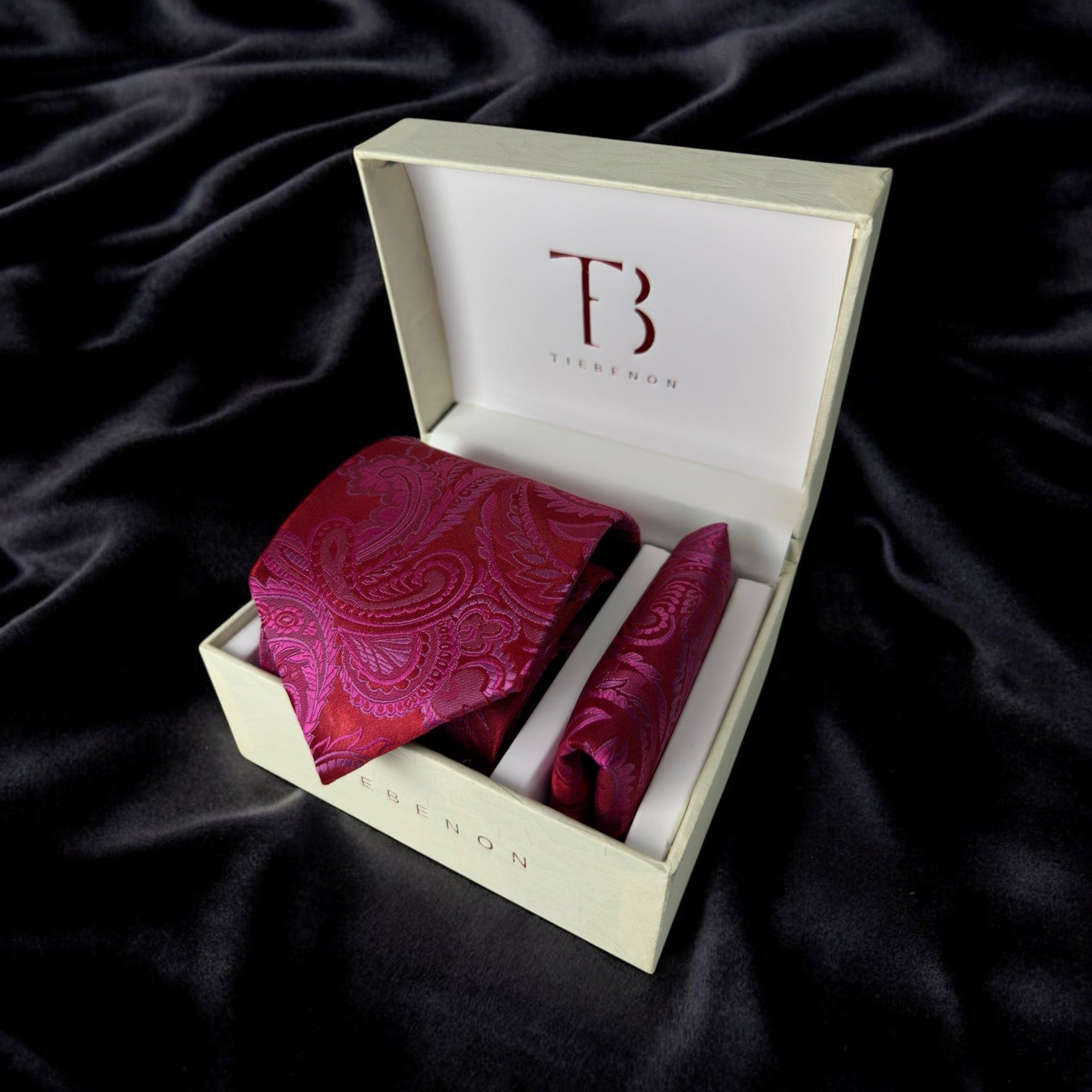 A hot pink paisley tie with a matching pocket square, presented in an open white box with a luxurious interior lining, resting on a plush black surface.