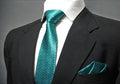 A turquoise geometric patterned tie and pocket square displayed on a mannequin wearing a black suit.