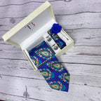 A violet blue traditional paisley silk tie with a floral pattern, displayed in an open white box with a branded logo on the top and a blue flower pin on the lapel, placed on a wooden surface.