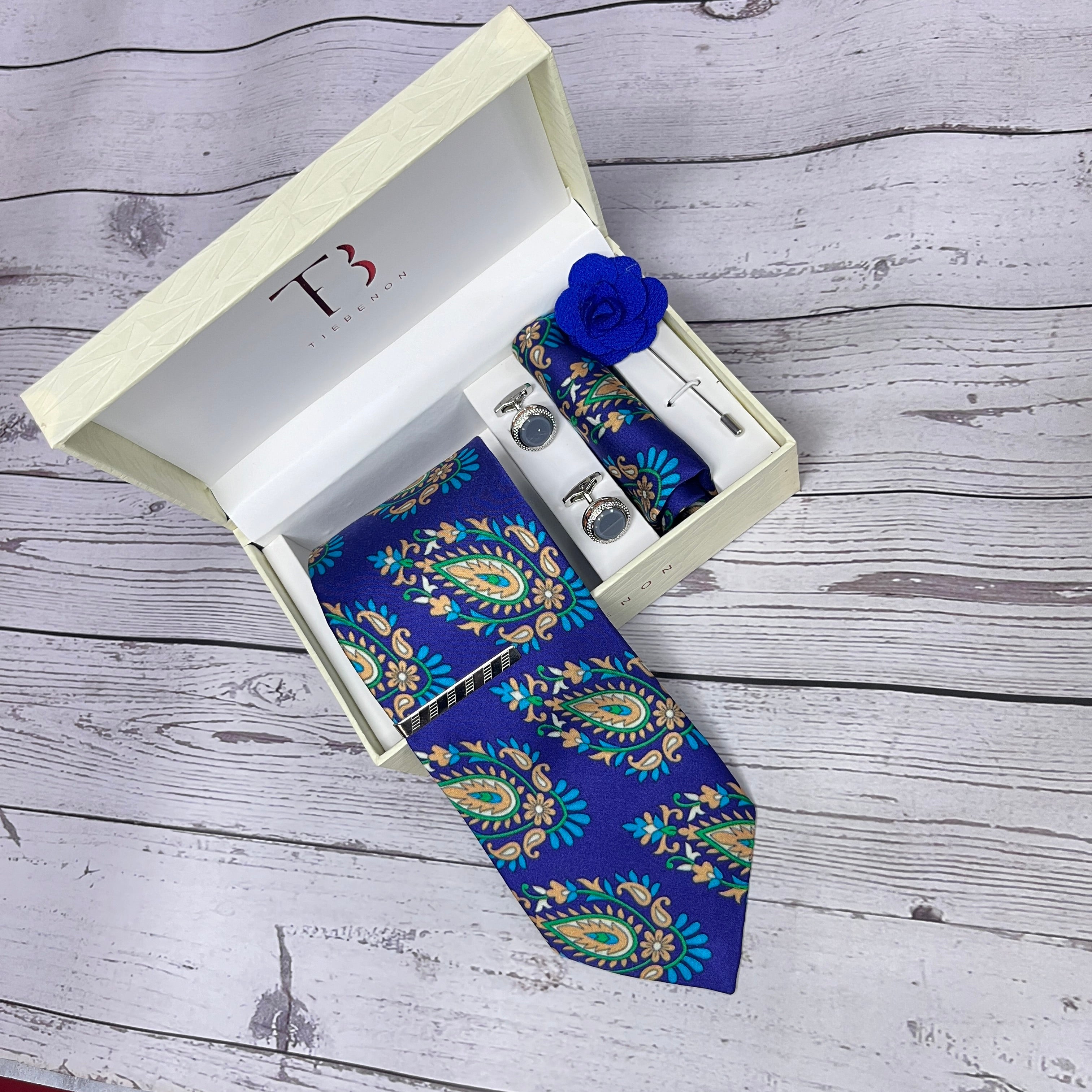 A violet blue traditional paisley silk tie with a floral pattern, displayed in an open white box with a branded logo on the top and a blue flower pin on the lapel, placed on a wooden surface.