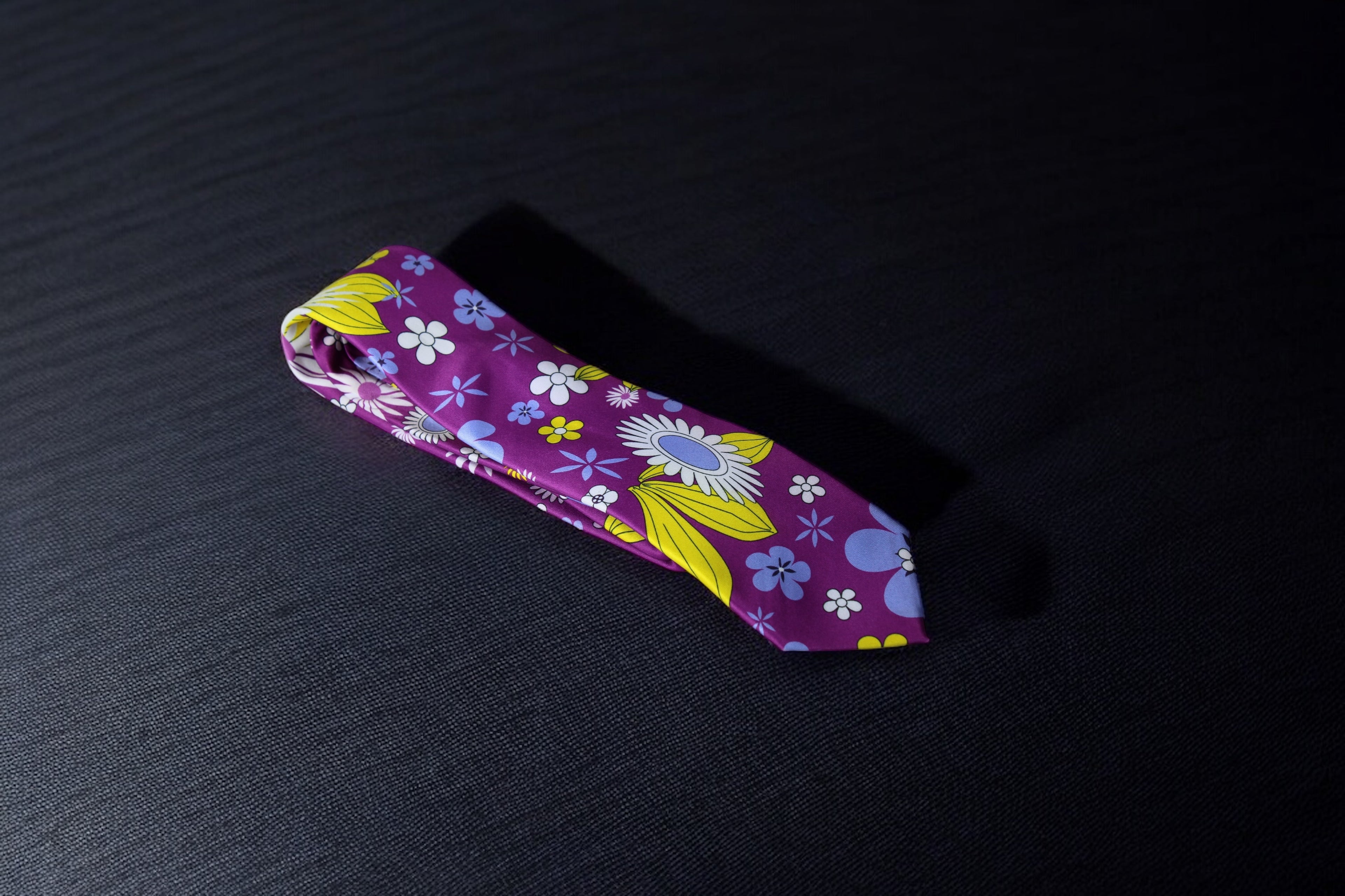 A purple necktie with a whimsical floral pattern laid out flat on a dark background.